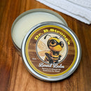 An open tin of NULU Inc.’s Dr. B.Strong’s Cutting Board Balm, featuring a cartoon bee on the lid, rests on a wooden surface. The white balm inside is formulated with beeswax and mineral oil, specially designed to provide superior protection for wooden cutting boards.