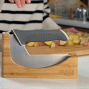 In the background, a person slices potatoes on a cutting board. In the foreground, the NULU Knife and Dock Bundle from NULU Inc.—featuring an ergonomic kitchen knife with a gray handle—rests on its wooden stand labeled “NULU,” offering convenient countertop storage.
