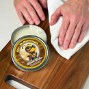 A tin of Dr. B.Strong’s Cutting Board Balm by NULU Inc. sits open on a wooden board, while hands use a white cloth to polish the surface—demonstrating the nourishing power of beeswax and mineral oil for optimal wood protection, just like an expert cutting board balm.