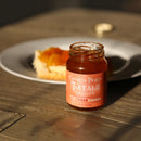 A jar of Powerful Pepper Company’s Powerful Pepper Peach Fatalii Jam rests on a wooden table, made with the finest Georgia peaches and fiery Fatalii peppers; in the blurred background, a biscuit topped with jam sits on a white plate.