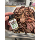 A hand holds a BuffalowBBQ Signature Blend BBQ Rub jar in front of a metal tray filled with sliced, cooked beef.