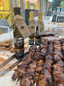 Grilled meat skewers on a wooden board are arranged beside bottles of Georgetown Olive Oil Co. Black Truffle & Garlic Oil and stacks of sliced bread, all set on an outdoor table with city buildings in the background.