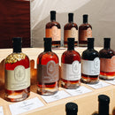Under The Bark Syrup Co Cardamom Maple Syrup bottles, labeled with flavors like cardamom infused, coffee, and smoked, are displayed on wooden shelves with tasting cards in front for easy sampling.