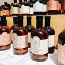 Bottles of Under The Bark Syrup Co Vanilla Maple Syrup are displayed on tables and wooden shelves, each featuring a leaf design label and the Under The Bark Syrup Co name.