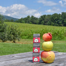 Three apples and three stacked tins of Spicewalla Apple Pie Spice by Spicewalla, a perfect seasonal blend for fall baking, rest on a wooden table outdoors with a grassy field, trees, and blue sky in the background.