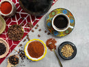 A cup of black coffee rests on a vibrant saucer, encircled by coffee beans, ground and whole spices, and a red patterned cloth atop a gray surface—evoking the rich aromas of World Spice Kaffe Baharat by World Spice and celebrating the essence of Middle Eastern flavors.