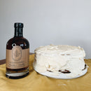 A bottle of Under The Bark Syrup Co Bourbon + Vanilla Maple Syrup is next to a white-frosted homemade cake on a white plate, both set on a yellow cloth against a plain white background.