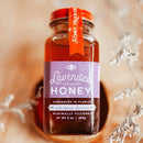 A jar of Honey Truck Co Lavender Infused Honey, featuring a purple label, rests on a wooden surface amid delicate dried flowers. The label notes it is harvested in Florida with French lavender, creating a soothing, lavender-infused honey from Honey Truck Co.