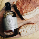A bottle of Under The Bark Syrup Co Cardamom Maple Syrup, with visible cardamom pods, rests on a wooden board beside a rustic loaf of bread with one slice cut.