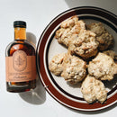 A bottle of Under The Bark Syrup Co Habanero Maple Syrup is placed beside six homemade biscuits on a white surface, with sunlight casting shadows.