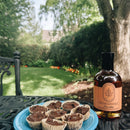 A bottle of Under The Bark Syrup Co Bourbon Maple Syrup and a plate of chocolate treats sit on a blue metal table in a garden with flowers and green trees in the background on a sunny day.