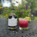 A glass of purple cocktail with a sugared rim and citrus slice sits beside a bottle of Under The Bark Syrup Co Blueberry Maple Syrup, its rich flavor showcased on a patterned outdoor table among purple and yellow flowers.