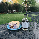 A slice of blueberry cheesecake on a blue plate and a bottle of Under The Bark Syrup Co Blueberry Maple Syrup rest on an ornate metal table outdoors, with grass, trees, and garden benches in the background.