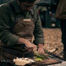 A person wearing a brown apron finely chops green herbs on a wooden cutting board outdoors, using the Messermeister Iron & Resin Overland Chef 8″ Camp Knife by Messermeister, with cheese and a mushroom nearby.