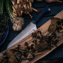 The Messermeister Iron & Resin Overland Chef 8″ Camp Knife, featuring a precision-forged blade, rests on a wooden board with grilled mushrooms, surrounded by fresh herbs, garlic, and raw mushrooms atop a dark tabletop.