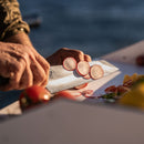 With sunlight illuminating their hands and the fresh vegetables, a person thinly slices a radish on a cutting board using the Messermeister Iron & Resin Overland Chef 8″ Camp Knife by Messermeister.