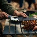 A person holds the Messermeister Iron & Resin Overland Chef 8″ Camp Knife in a leather sheath over a picnic table scattered with mushrooms and vegetables—an ideal companion for outdoor cooking adventures from Messermeister.