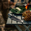 The Messermeister Iron & Resin Overland Chef 8″ Camp Knife rests in its sheath on a folding table beside a cluster of mushrooms and herbs, with blurred outdoor cooking gear and a campfire visible in the background.