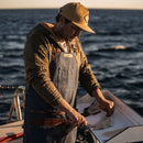 A person wearing a cap and apron is filleting fish on a boat at sea during sunset, using the Messermeister Iron & Resin Overland Chef 7″ Mid-Flex Fillet Knife, with ocean waves in the background.