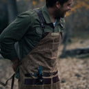 A man in a green shirt and brown apron stands outdoors, smiling as he adjusts his apron strap. Tucked into one of the apron’s pockets is a Messermeister Iron & Resin Overland Chef 7″ Mid-Flex Fillet Knife, featuring a blue handle and crafted from high-carbon stainless steel—perfect for precise fish preparation. The background is softly blurred with autumn leaves scattered on the ground.