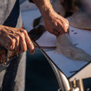 A person expertly fillets a fish on a white cutting board, sunlight highlighting their hands and the fish’s fresh flesh, using the Messermeister Iron & Resin Overland Chef 7″ Mid-Flex Fillet Knife by Messermeister.