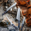 Four Messermeister kitchen knives, including the Iron & Resin Overland Chef 8″ Camp Knife—known for its precision-forged blade and suitability for outdoor cooking—are displayed among rocks, seashells, cooked crabs on wooden boards, and a blue crab set on newspaper. Together they create a seafood preparation scene filled with diverse textures and natural elements.