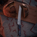 A loaf of rustic bread, partially sliced with the Messermeister Iron & Resin Overland Chef 8″ Bread Knife, sits on a wooden board atop a patterned cloth.