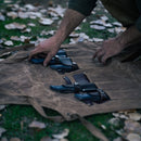 A person arranges knives—including the Messermeister Iron & Resin Overland Chef 8″ Bread Knife—in leather sheaths on a brown canvas roll, placed on grass with scattered dry leaves, ready for outdoor cooking.