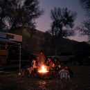 Five people sit around a campfire at dusk, surrounded by trees, a trailer, and parked vehicles. Camping gear and string lights are visible, creating a cozy outdoor atmosphere as someone prepares for outdoor cooking with the Messermeister Iron & Resin Overland Chef 8″ Bread Knife resting nearby.