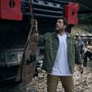 A man in a green jacket holds up a brown utility apron near a truck equipped with fuel canisters. People sit outdoors among fallen leaves in the background, while the Messermeister Iron & Resin Overland Chef 9″ Carving Knife by Messermeister peeks out from one of the apron’s pockets.