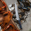 A Messermeister Iron & Resin Overland Chef 9″ Carving Knife lies on sand and rocks beside seashells, cooked crabs on a wooden board, and a live blue crab.