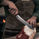 Wearing a brown apron, a person uses the Messermeister Iron & Resin Overland Chef 9″ Carving Knife to slice raw meat on a wooden surface, with the focus on their hands and the meat.