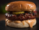 Close-up of a juicy beef burger with pickles and creamy sauce, topped with Terrapin Ridge Farms Hot Pepper Bacon Jam 1 Gallon Jug for sweet heat, all on a golden bun. Brand: Terrapin Ridge Farms. Blurred background.