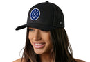 A woman with long brown hair smiles while wearing the Haka Hat Indianapolis Trucker Hat—a black IND X Football snapback with a mesh back and round blue-and-white logo—against a plain white background.