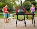 A red outdoor wood-fired pizza oven on a wheeled stand is paired with the WPPO LLC Direct Infinity Side Table Short Side 75cm, which fits Infinity 50 and 66 models and includes firewood storage, as a family enjoys a backyard gathering in the background.