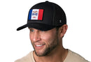 A man with light facial hair smiles while wearing the Haka Hat Iowa Flag Trucker Hat, a black mesh snapback by Haka Hat featuring an Iowa state flag patch. He pairs it with a light shirt against a white background.