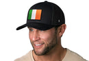 A smiling man with a trimmed beard wears the Haka Hat Ireland Flag Trucker Hat, a black Irish snapback by Haka Hat featuring an embroidered green, white, and orange flag patch. He’s in a light beige shirt against a white background.