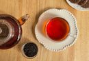 A glass teacup filled with World Spice Irish Breakfast tea on a white plate, a glass teapot, a small bowl of World Spice loose black tea leaves, and a plate of chocolate cookies on a wooden table.