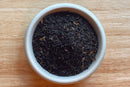 A small white bowl filled with loose World Spice Irish Breakfast tea leaves from World Spice sits on a light wooden surface.