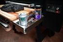A Pimp My Grill® Tactical Stainless Folding Front Shelf for Traeger Pro 22 / Pro 575 / Redland / Ironwood 650 attached to a pellet grill, serving as a prep station with a beverage, thermometer, rub, and a brisket on a cutting board.