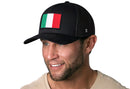 A man wearing a Haka Hat Italian Flag Trucker Hat | Black Italy Snapback with a bold embroidered flag patch smiles to the side. He has short brown hair, a trimmed beard, and wears a beige shirt.