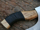 A close-up of the Texan Knives Ax Pizza Cutter—custom handcrafted with a J2 steel blade and an engraved, curved ash wood handle partially wrapped in black cord—rests on a textured, bark-like brown surface.