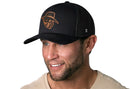 A man with a trimmed beard and mustache smiles while looking slightly to the side against a white background, wearing a beige t-shirt and the Haka Hat JBR Logo Black Poly Leather Snapback by Haka Hat.