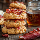 A stack of cookies topped with crispy bacon sits beside syrup, all seasoned with SALTOPIA Infused Sea Salts' Bacon Maple Seasoning: JURASSIC PORK, adding a sweet-smoky flavor. More cookies and bacon are visible in the background.