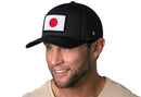 A smiling man wears the Haka Hat Japanese Flag Trucker Hat | Black Japan Snapback by Haka Hat, featuring an embroidered flag patch on the front and a beige shirt, set against a white background.