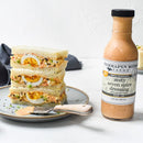 A plate with a stacked egg salad sandwich garnished with herbs sits beside an open jar of Terrapin Ridge Farms Zesty Seven Spice Dressing. A knife with the dressing rests on the plate, ready for spreading.