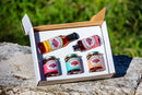 An open gift box labeled Brushfire Farms El Jefe Bundle sits on a rock in a grassy, blurred background, featuring two bottles of Brushfire Farms hot sauce and three jars of Three Amigos Jam set, all with vibrant labels.