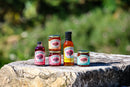 Five Brushfire Farms products (jars, bottles) are displayed on a large rock with a blurred green natural background. Among them, the popular El Jefe Bundle stands out, each label featuring Brushfire Farms’ distinctive red and white design. Perfect as a gift box or for those who love pepper jam, these jars showcase vibrant flavors inspired by nature’s bounty.