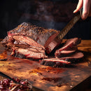 A person slices juicy, smoked brisket on a wooden board using the SpitJack Deluxe Brisket Slicing Knife – 11 Inch Blade, Competition-Chef Series. The meat is tender with a dark, seasoned crust, and steam rises from the fresh, hot slices.