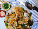 A plate of roasted chicken with herbs and vegetables sits on a striped cloth next to the SpitJack 8 Inch Chef Knife and 11 Inch Sharpening Hone Combo, along with a bowl of chopped parsley and a small dish of sesame seeds.